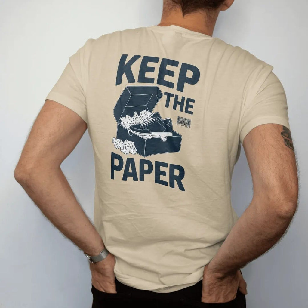 Man wearing a Sandstone tee, seen from the back: an open shoebox overflowing with crumpled tissue paper and a nested sneaker, under Keep The and above Tissue Paper, as a two-ink streetwear graphic in navy and orange.