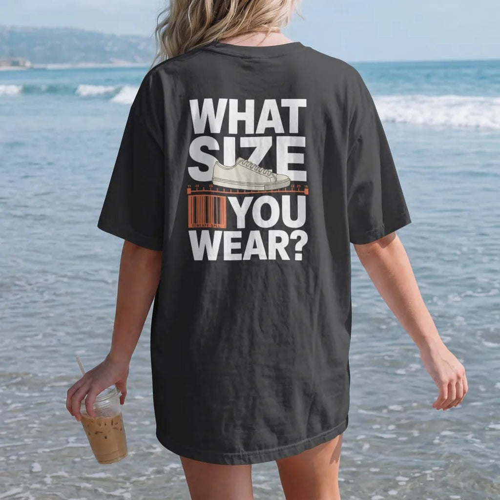 Woman on the beach in a Black tee, back view: a shoe-store foot measuring device holding a sneaker under What Size and You Wear?, as a two-ink streetwear graphic in off-white and orange.