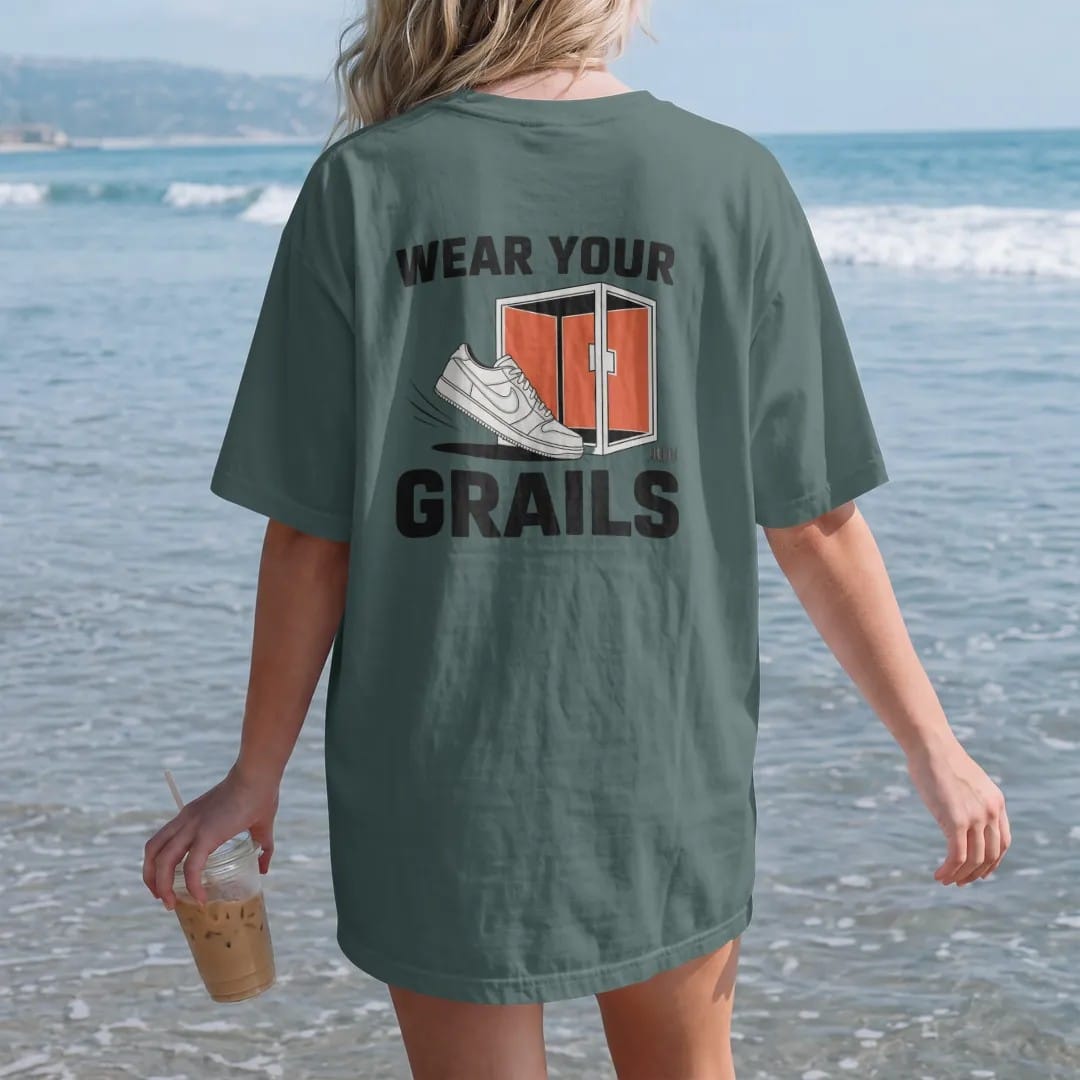 Woman on the beach in a Blue Spruce tee, back view: a sneaker stepping out of an open glass display case under Wear Your and a huge Grails, as a two-ink streetwear graphic in off-white and orange.