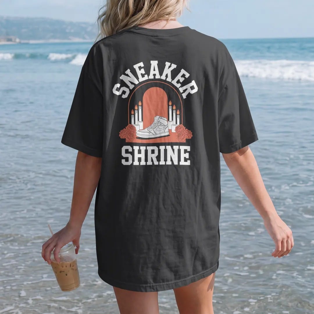 Woman on the beach in a Black tee, back view: a candlelit altar with roses and a single sneaker under a glowing arch, with Sneaker above and Shrine below, as a two-ink streetwear graphic in off-white and orange.