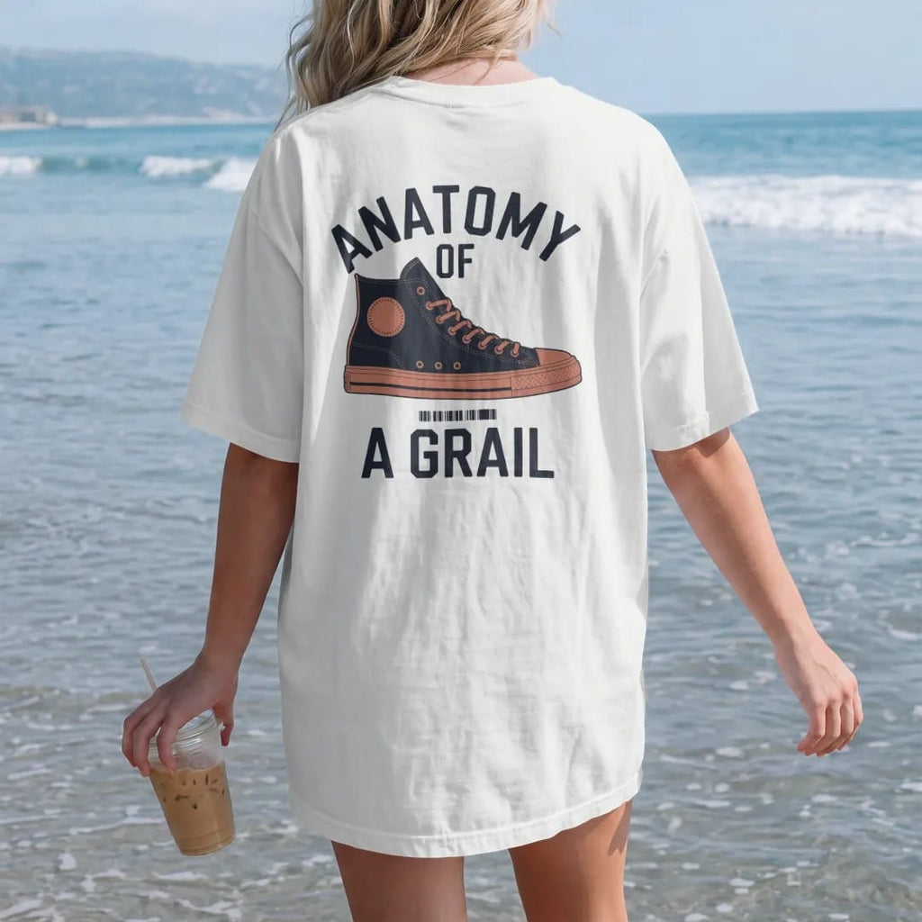 Woman on the beach in a White tee, back view: an exploded diagram of a high-top sneaker with leader lines under Anatomy Of and above A Grail, as a two-ink streetwear graphic in navy and orange.