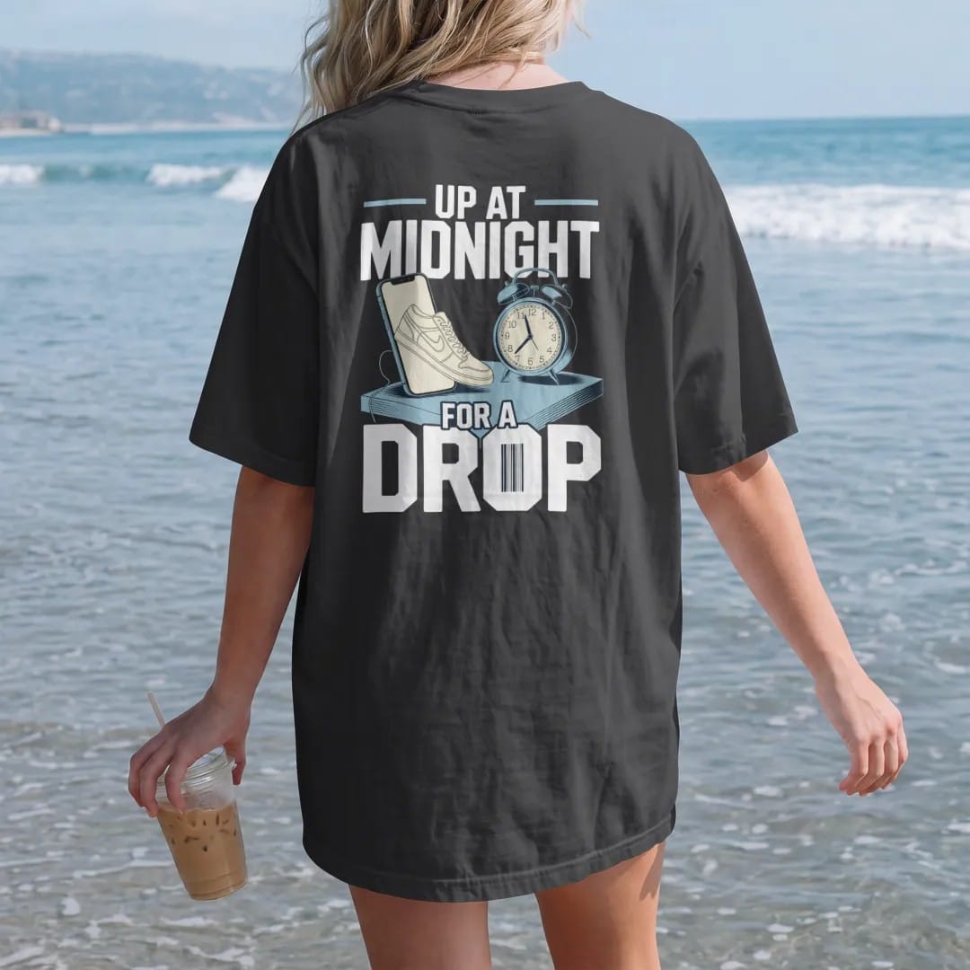 Woman on the beach in a Black tee, back view: a glowing phone with a sneaker on screen beside an alarm clock at midnight, under Up At Midnight and For A Drop, as a two-ink streetwear graphic in off-white and icy blue.
