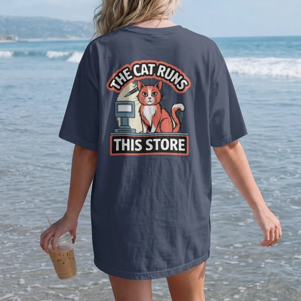 Woman on the beach in a Navy tee, back view: an orange cat behind a register with a pen behind its ear, framed by The Cat Runs and This Store, in halftone comic-pop red, yellow and cream.