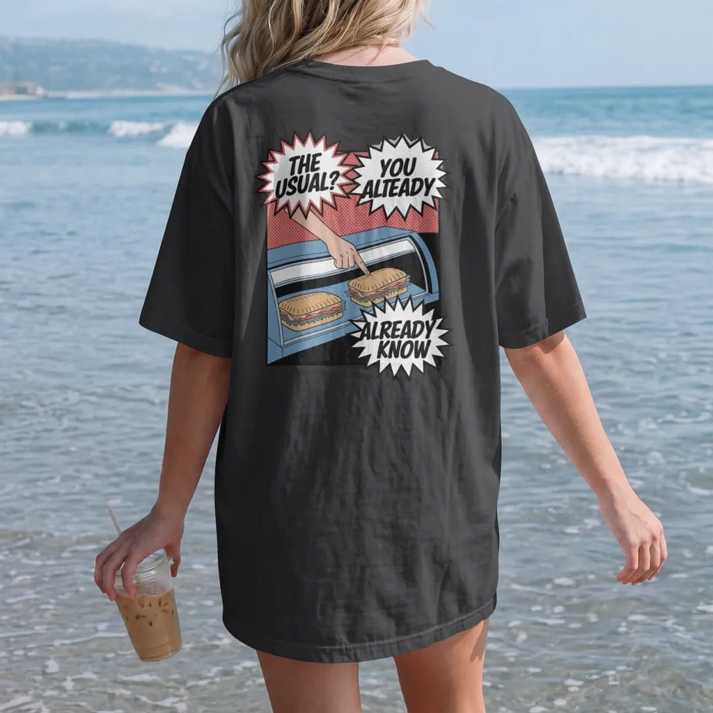 Woman on the beach in a Black tee, back view: a deli counter with a hand pointing at a wrapped sandwich and speech bubbles reading The Usual? and You Already Know, in halftone comic-pop red, yellow and cream.