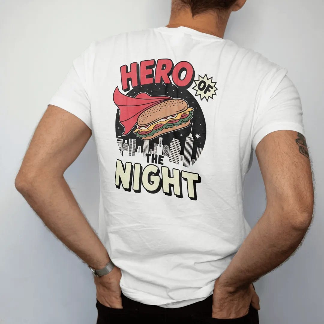 Man wearing a White tee, seen from the back: a caped hero sandwich flying over a night skyline under comic Hero Of and The Night letters, in halftone comic-pop red, yellow and cream.