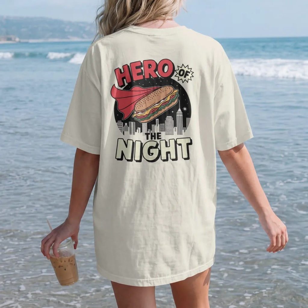 Woman on the beach in a Ivory tee, back view: a caped hero sandwich flying over a night skyline under comic Hero Of and The Night letters, in halftone comic-pop red, yellow and cream.