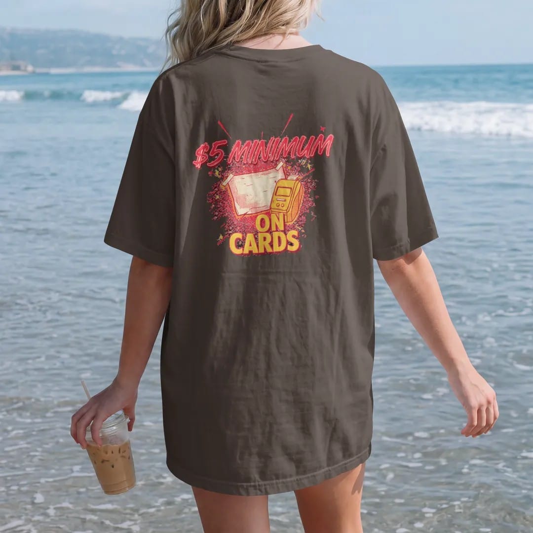 Woman on the beach in a Espresso tee, back view: a taped handwritten sign on a register beside a card reader and gum reading $5 Minimum On Cards, in halftone comic-pop red, yellow and cream.