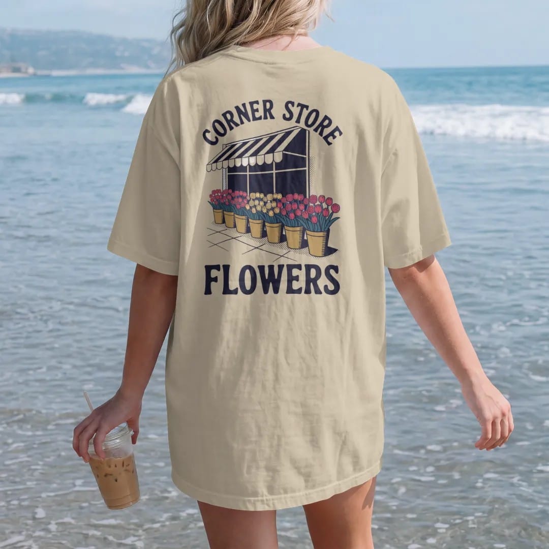 Woman on the beach in a Sandstone tee, back view: buckets of tulips, roses and carnations under a striped awning with Corner Store arched above and Flowers below, in halftone comic-pop red, mustard and navy.