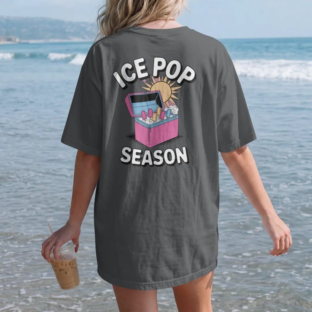 Woman on the beach in a Graphite tee, back view: an open ice-pop freezer with colorful popsicles and frosty mist under a big sun, with melting Ice Pop and Season, in halftone comic-pop pink, yellow and cream.