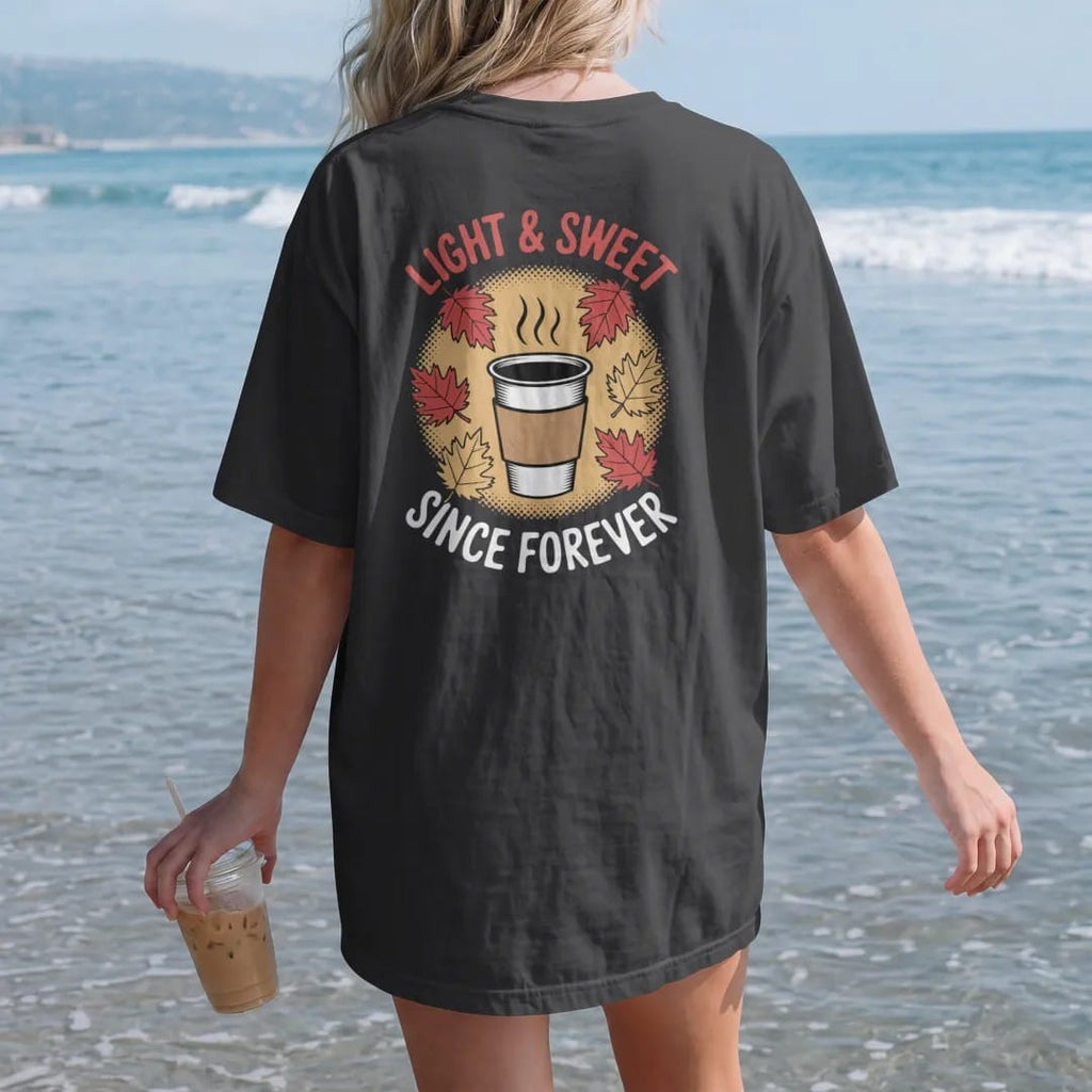 Woman on the beach in a Black tee, back view: a steaming paper coffee cup with swirling autumn leaves under Light & Sweet script and Since Forever, in halftone comic-pop red, yellow and cream.