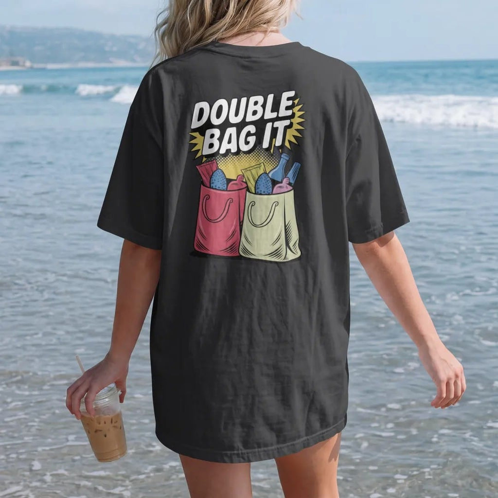 Woman on the beach in a Black tee, back view: two plastic bags nested and stuffed with groceries and a bottle peeking out, under Double Bag It, in halftone comic-pop red, yellow and cream.