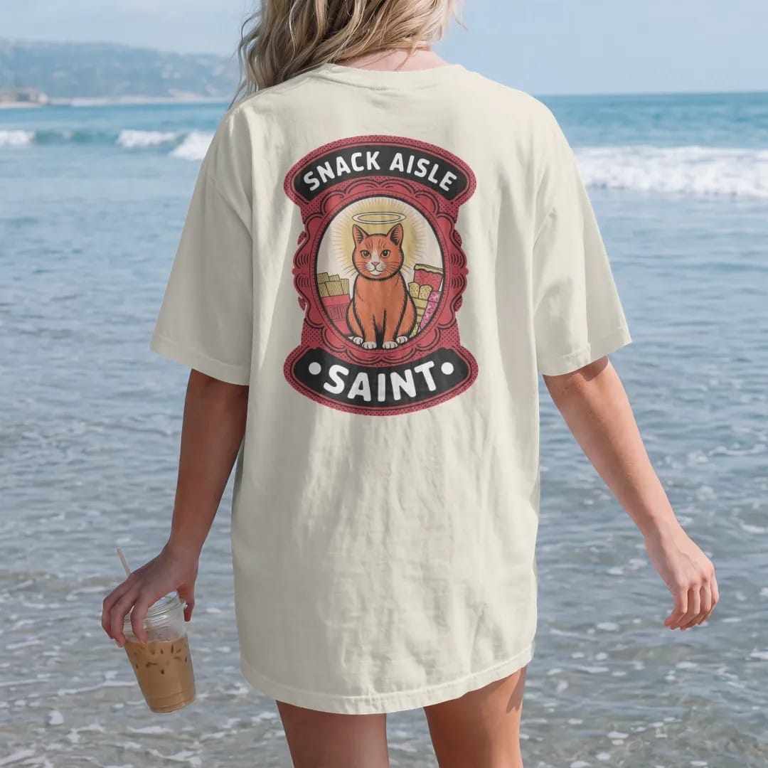 Woman on the beach in a Ivory tee, back view: a prayer-candle label of a haloed orange cat surrounded by chips and candy, titled Snack Aisle Saint, in halftone comic-pop red, yellow and cream.