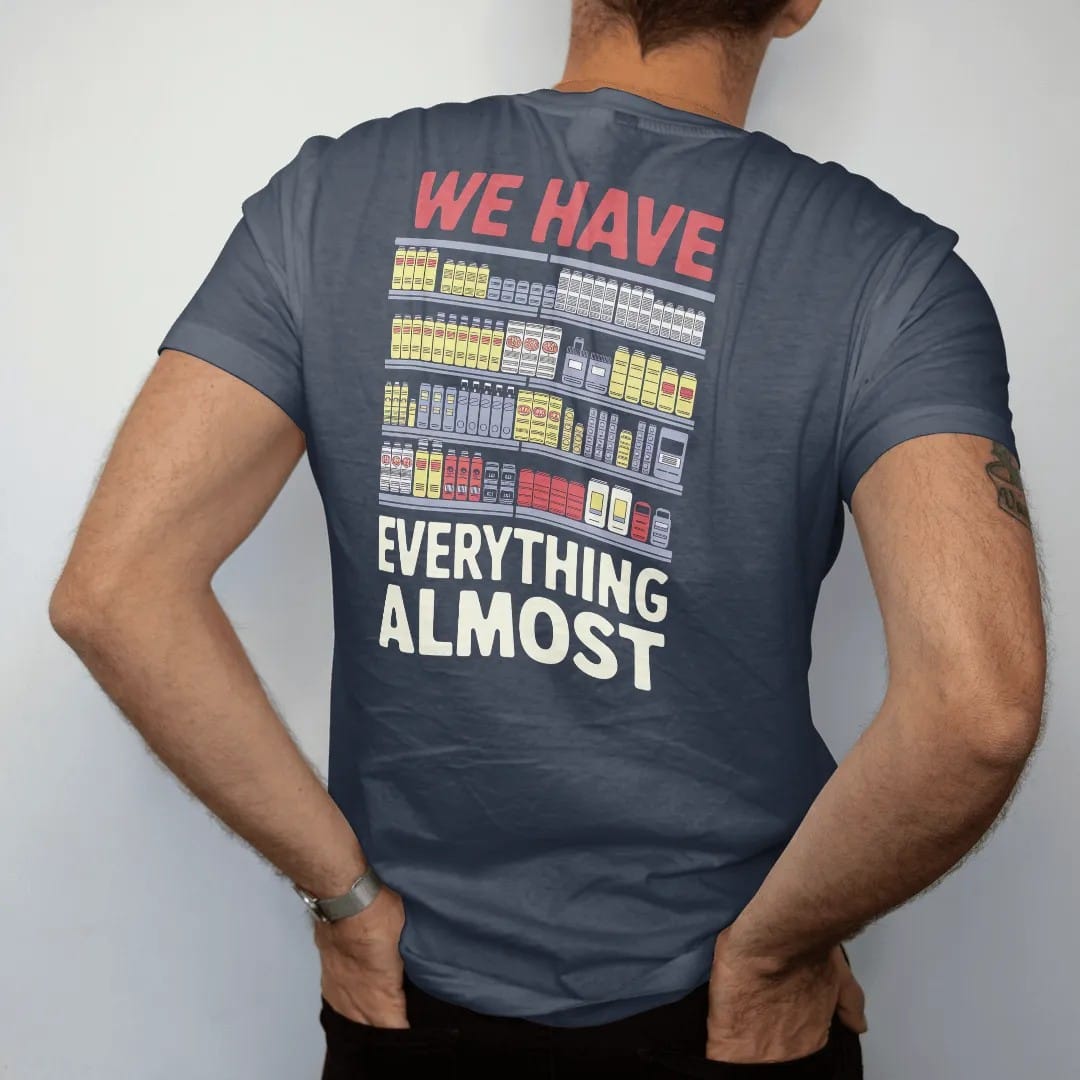 Man wearing a Midnight tee, seen from the back: crowded shelves of cans, batteries, cereal, chargers and an umbrella under We Have Everything and a tiny Almost, in halftone comic-pop red, yellow and cream.
