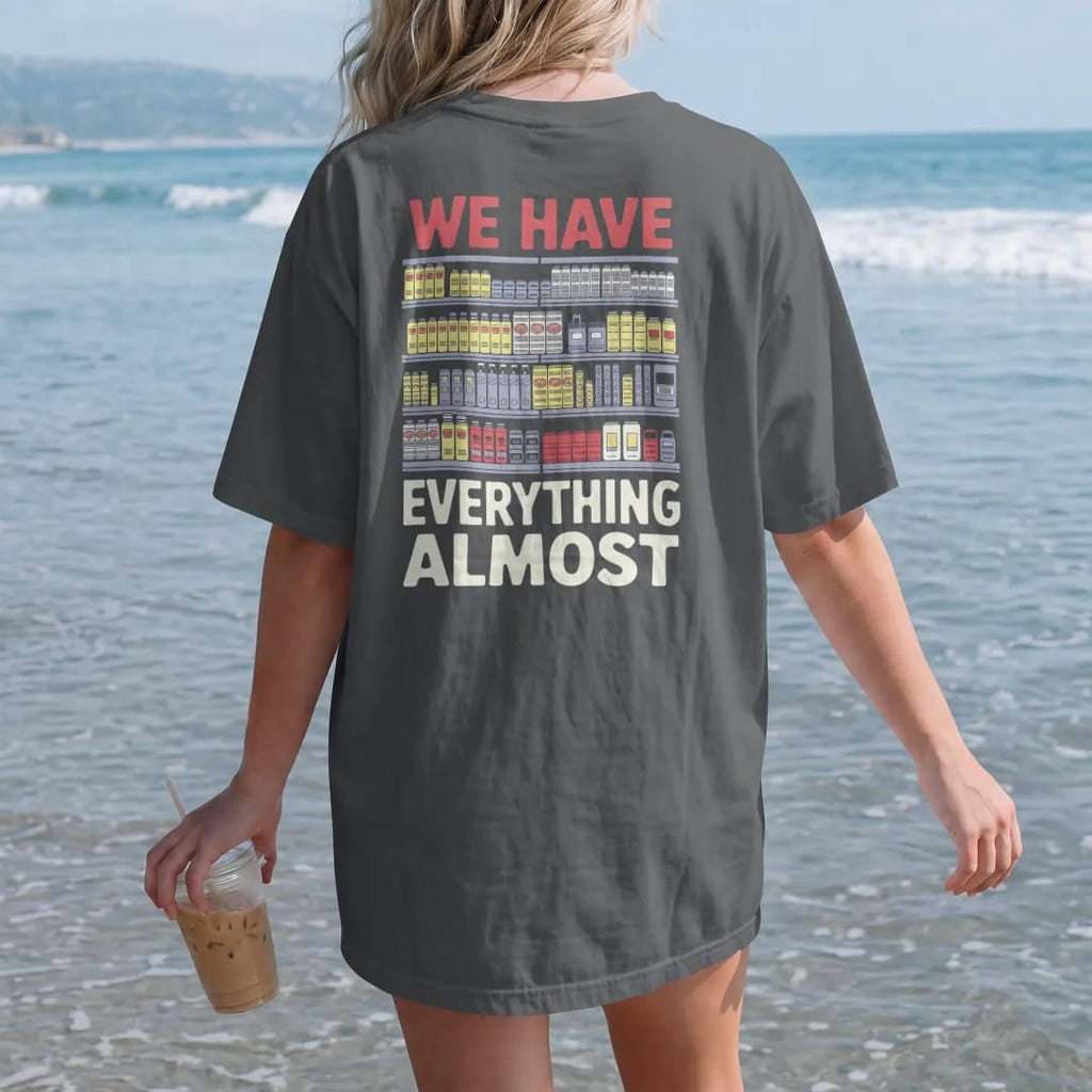 Woman on the beach in a Graphite tee, back view: crowded shelves of cans, batteries, cereal, chargers and an umbrella under We Have Everything and a tiny Almost, in halftone comic-pop red, yellow and cream.