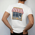 Man wearing a White tee, seen from the back: a striped scalloped awning painted with Grocery, Deli and Candy over a cat on a windowsill, in halftone comic-pop red, mustard and navy.
