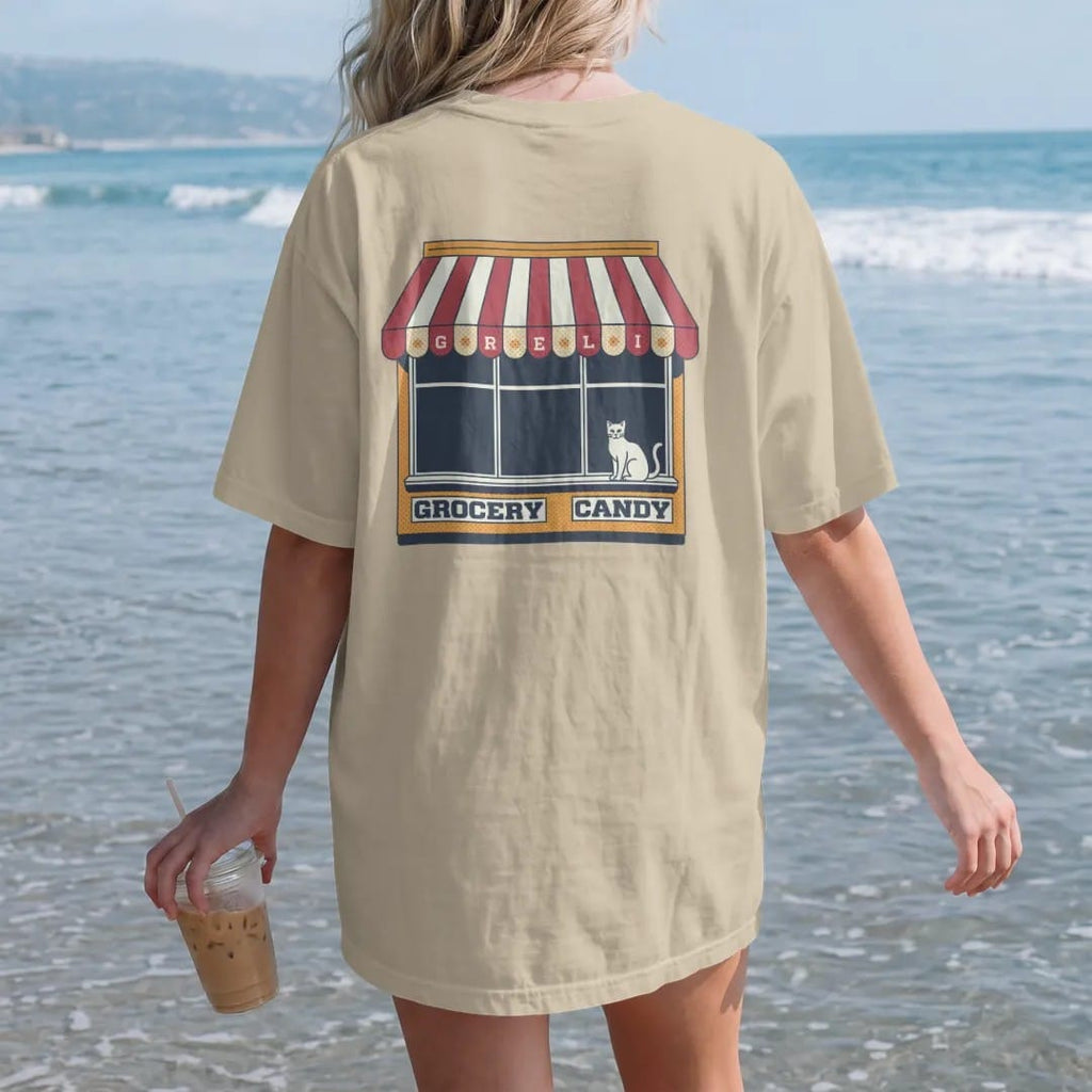 Woman on the beach in a Sandstone tee, back view: a striped scalloped awning painted with Grocery, Deli and Candy over a cat on a windowsill, in halftone comic-pop red, mustard and navy.