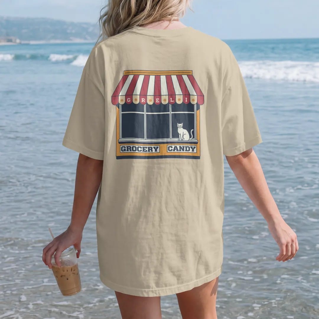 Woman on the beach in a Sandstone tee, back view: a striped scalloped awning painted with Grocery, Deli and Candy over a cat on a windowsill, in halftone comic-pop red, mustard and navy.