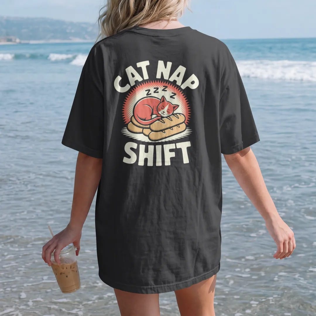 Woman on the beach in a Black tee, back view: an orange cat asleep on bread loaves with floating z's under Cat Nap and Shift, in halftone comic-pop red, yellow and cream.