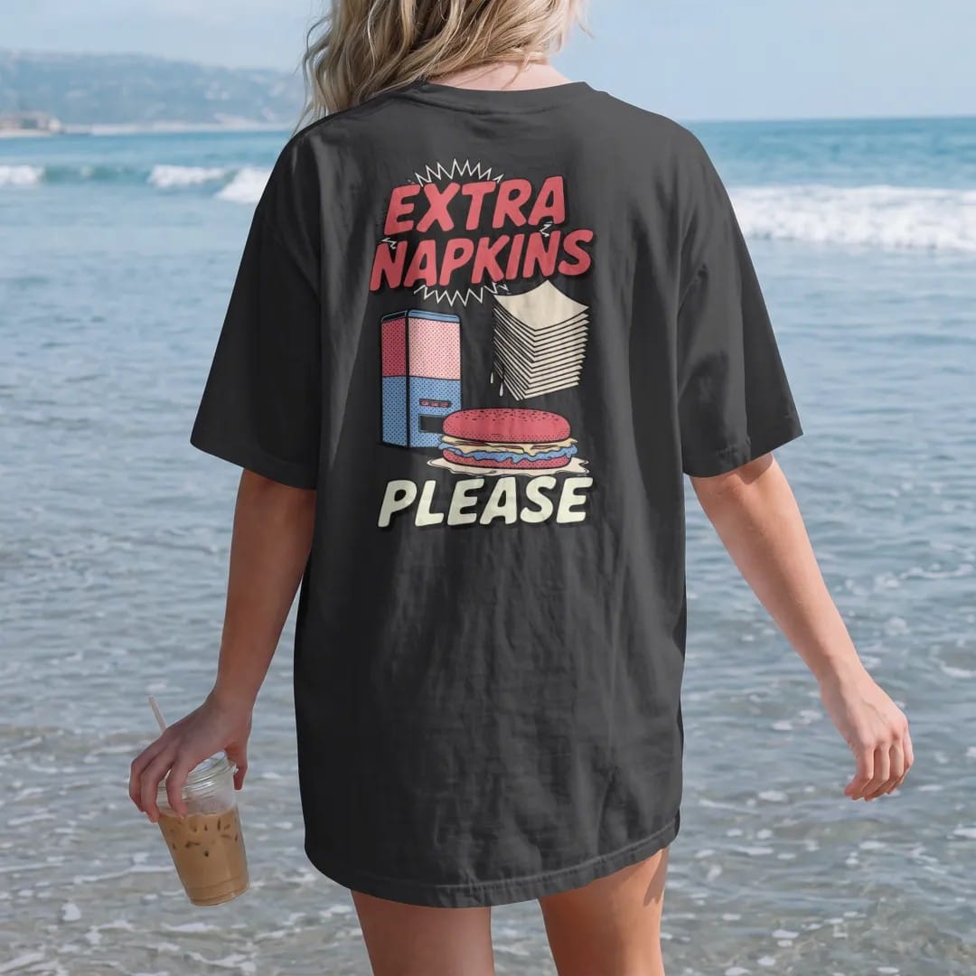 Woman on the beach in a Black tee, back view: a stack of napkins flying from a dispenser beside a dripping sandwich under Extra Napkins and Please, in halftone comic-pop red, yellow and cream.