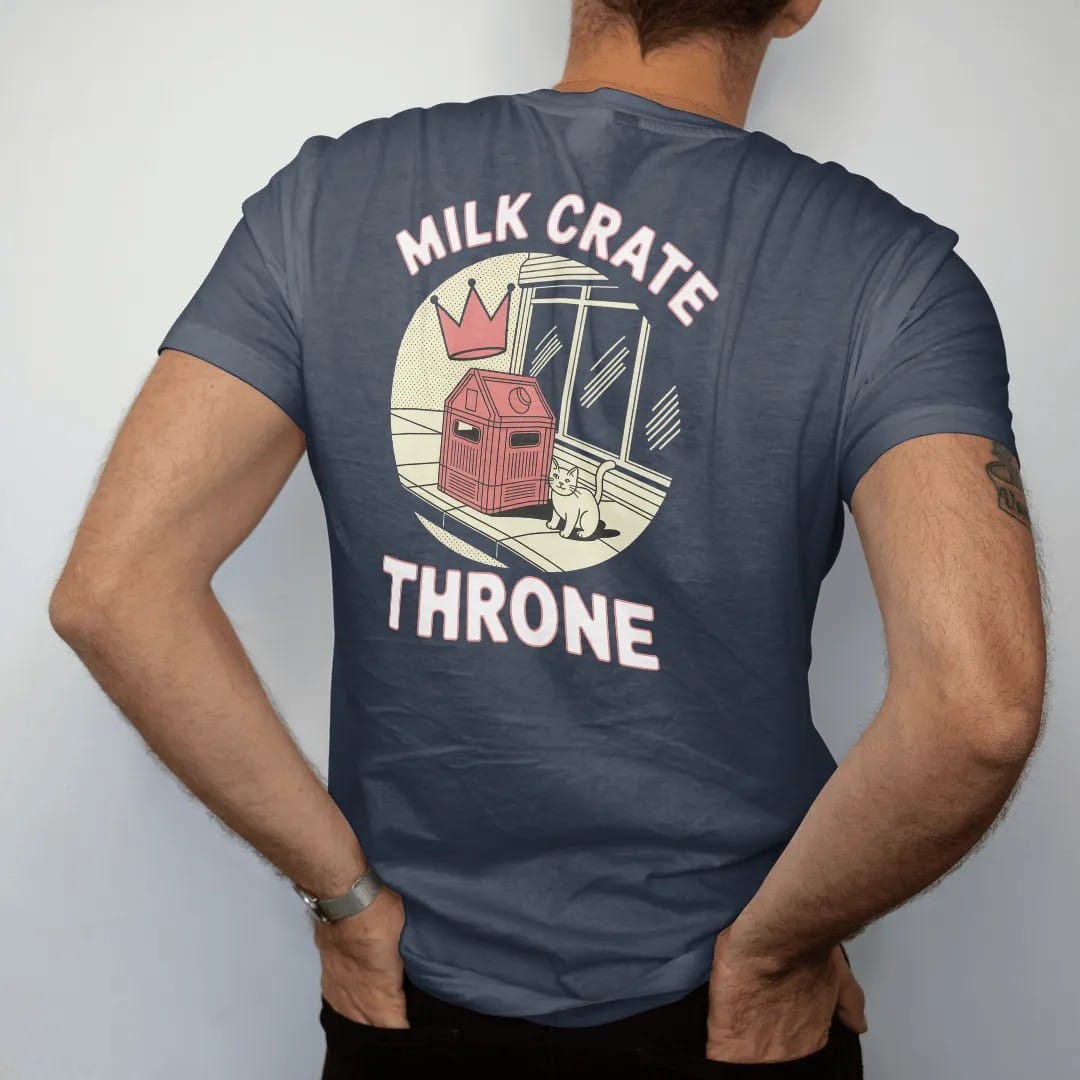 Man wearing a Midnight tee, seen from the back: a milk crate on the sidewalk with a floating crown and a cat beside it under Milk Crate and Throne, in halftone comic-pop red, yellow and cream.