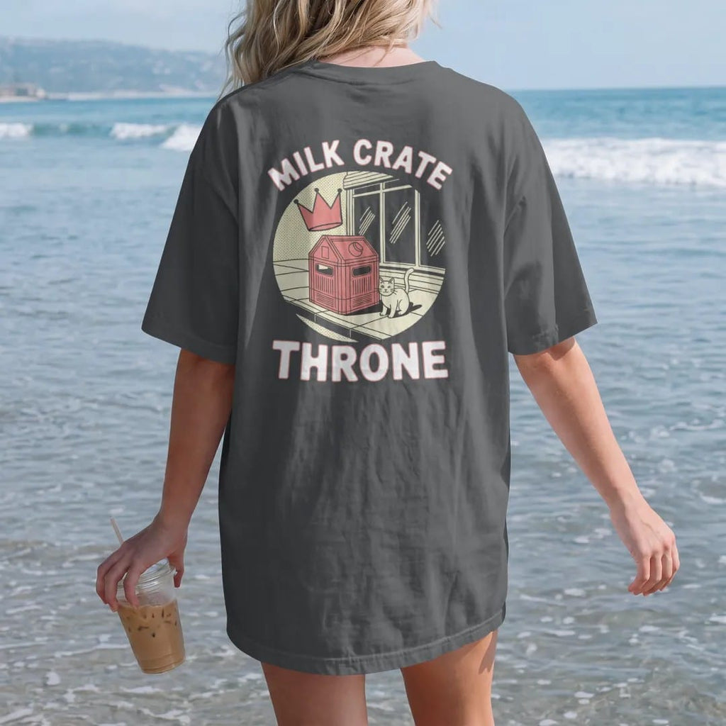 Woman on the beach in a Graphite tee, back view: a milk crate on the sidewalk with a floating crown and a cat beside it under Milk Crate and Throne, in halftone comic-pop red, yellow and cream.