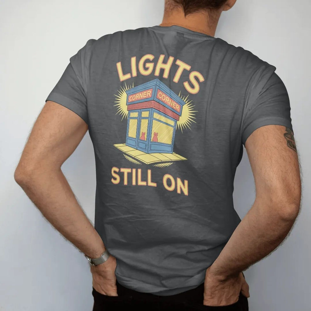 Man wearing a Graphite tee, seen from the back: a lone corner store glowing at the end of a dark street with a cat in the window, under neon Lights and Still On, in halftone comic-pop red, yellow and cream.