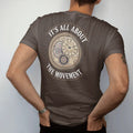 Man wearing a Espresso tee, seen from the back: a watch caseback revealing a mechanical movement with bridges, jewels and a rotor, under It's All About and The Movement, as a fine engraving in antique gold and cream.