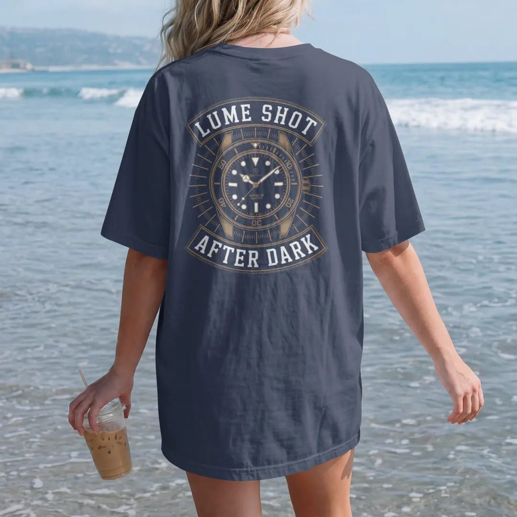 Woman on the beach in a Navy tee, back view: a dive watch dial with glowing hands and markers and faint rays, under glowing Lume Shot and After Dark, as a fine engraving in antique gold and cream.