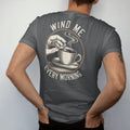 Man wearing a Graphite tee, seen from the back: fingers turning a watch winding crown with curved arrows and a steaming coffee cup, under Wind Me and Every Morning, as a fine engraving in antique gold and cream.