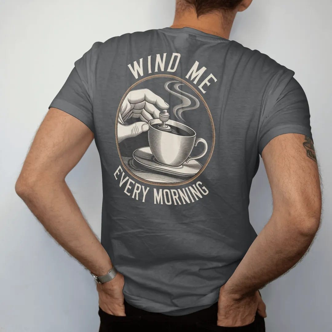 Man wearing a Graphite tee, seen from the back: fingers turning a watch winding crown with curved arrows and a steaming coffee cup, under Wind Me and Every Morning, as a fine engraving in antique gold and cream.
