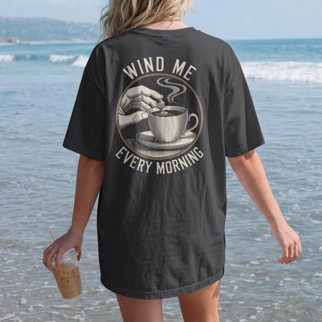 Woman on the beach in a Black tee, back view: fingers turning a watch winding crown with curved arrows and a steaming coffee cup, under Wind Me and Every Morning, as a fine engraving in antique gold and cream.