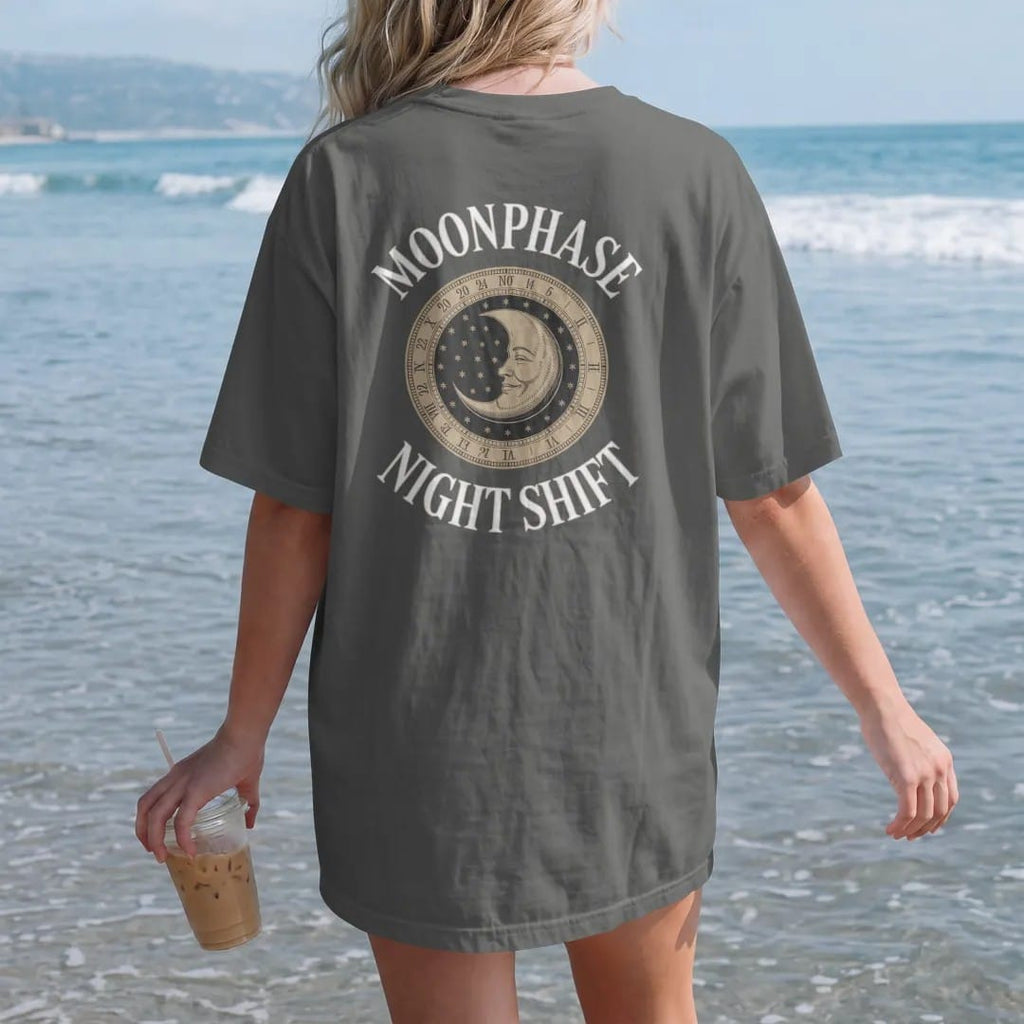Woman on the beach in a Pepper tee, back view: a moonphase window with an engraved smiling moon on a starry disc, under Moonphase and Night Shift, as a fine engraving in antique gold and cream.