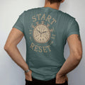 Man wearing a Blue Spruce tee, seen from the back: a chronograph watch with leader lines pointing to the pushers labeled Start, Stop and Reset, as a fine engraving in antique gold and cream.