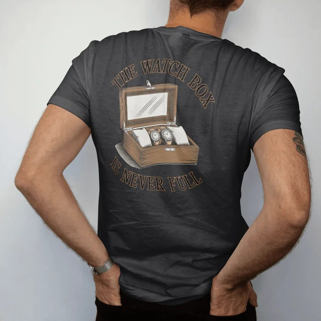 Man wearing a Black tee, seen from the back: an open glass-lidded watch box full of watches with one empty pillow highlighted, under The Watch Box and Is Never Full, as a fine engraving in antique gold and cream.