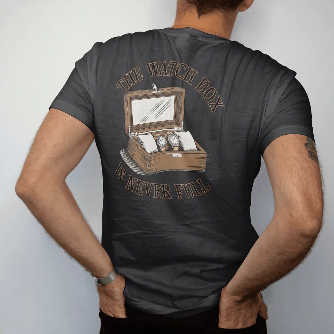 Man wearing a Black tee, seen from the back: an open glass-lidded watch box full of watches with one empty pillow highlighted, under The Watch Box and Is Never Full, as a fine engraving in antique gold and cream.