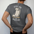 Man wearing a Graphite tee, seen from the back: a long waitlist scroll unrolling to a tiny watch and a cobweb, under Still On and The Waitlist, as a fine engraving in antique gold and cream.