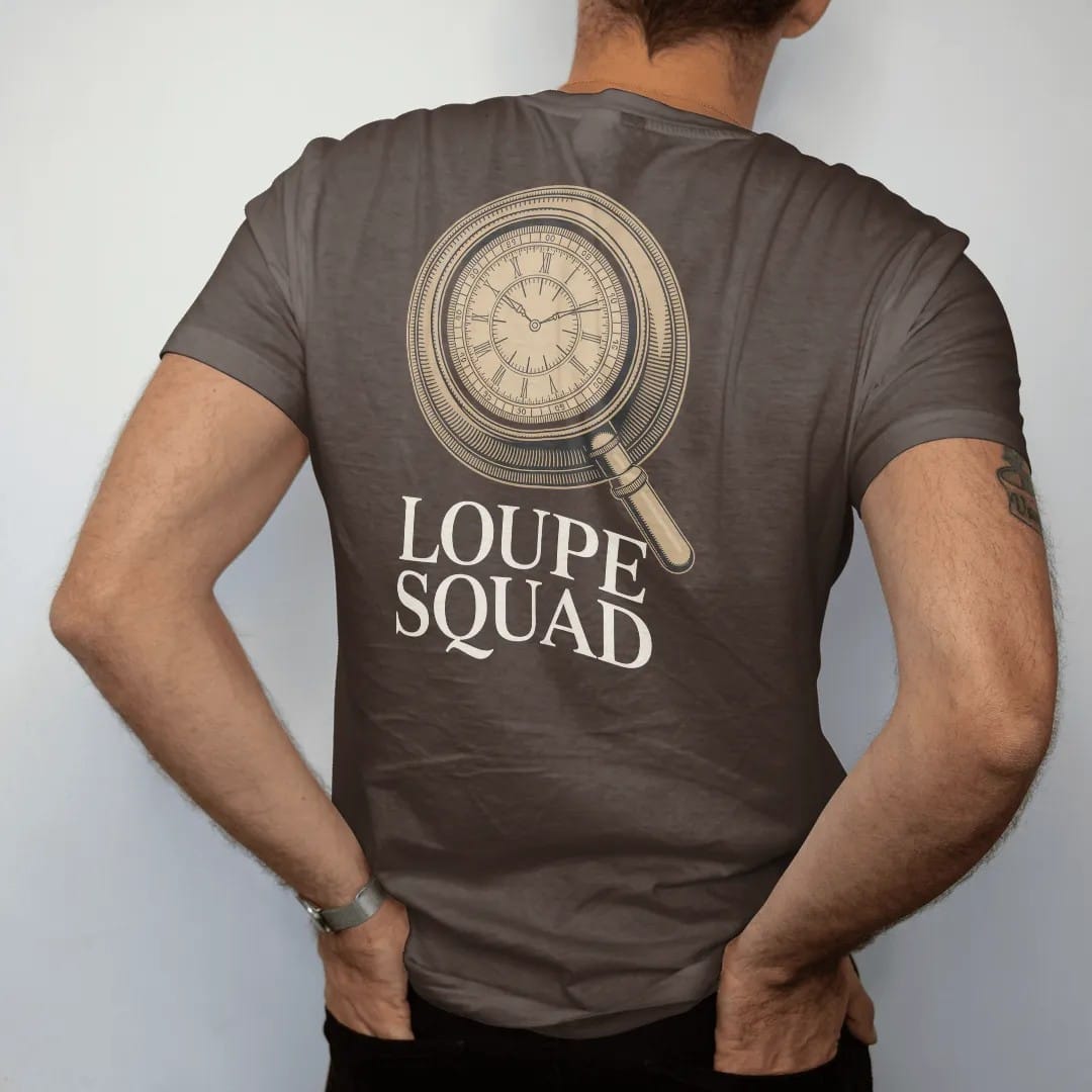 Man wearing a Espresso tee, seen from the back: a jeweler's loupe magnifying part of a watch dial with sharp detail inside the lens, above Loupe Squad, as a fine engraving in antique gold and cream.