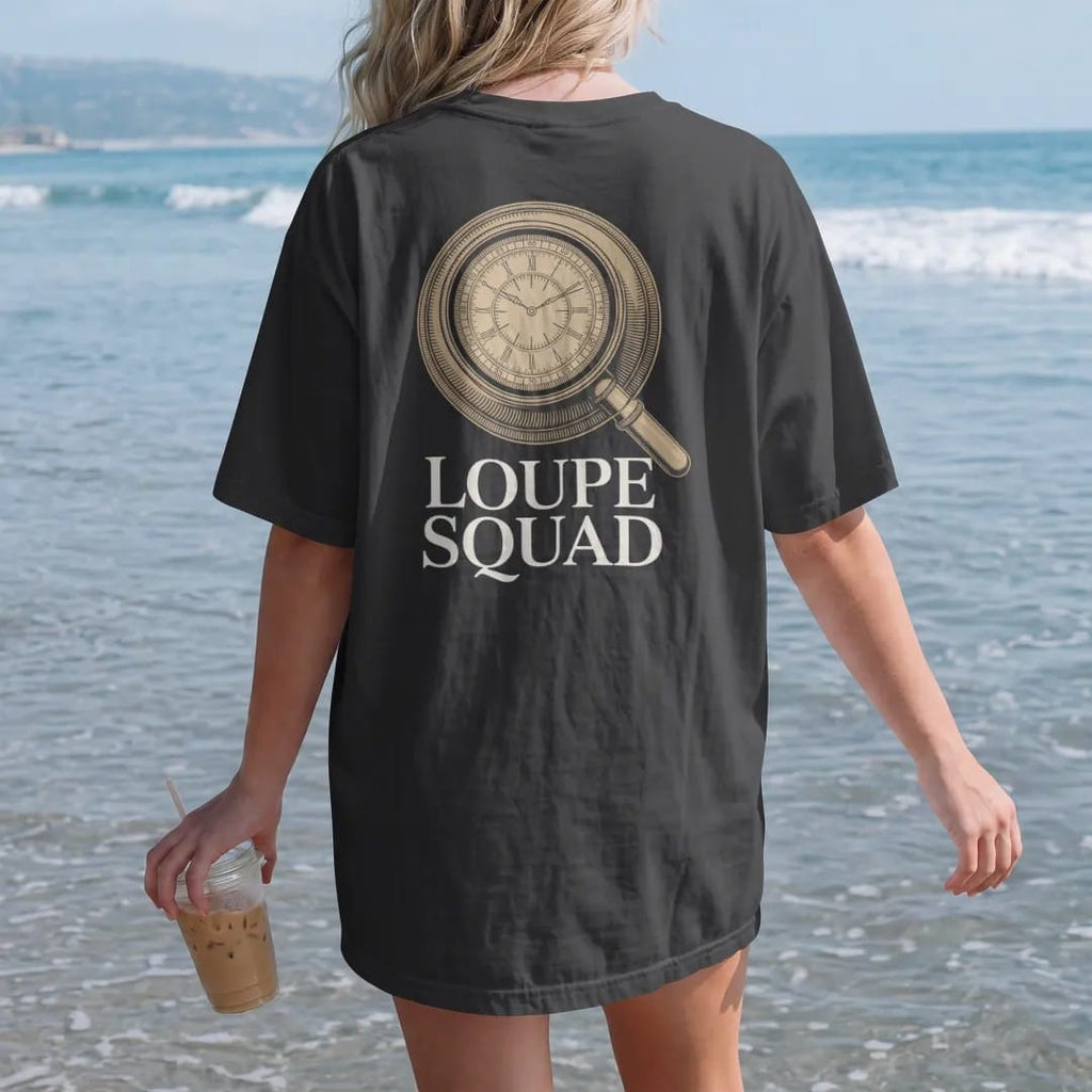 Woman on the beach in a Black tee, back view: a jeweler's loupe magnifying part of a watch dial with sharp detail inside the lens, above Loupe Squad, as a fine engraving in antique gold and cream.