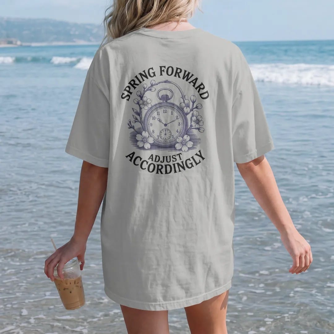 Woman on the beach in a Granite tee, back view: a watch with its crown pulled and hands advancing amid cherry blossoms, under Spring Forward and Adjust Accordingly, as a fine engraving in navy and antique brass.
