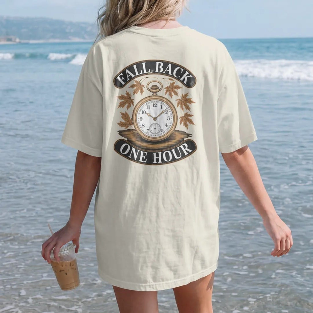 Woman on the beach in a Ivory tee, back view: a watch dial with rewinding hands surrounded by drifting maple leaves, under Fall Back and One Hour, as a fine engraving in antique gold and cream.