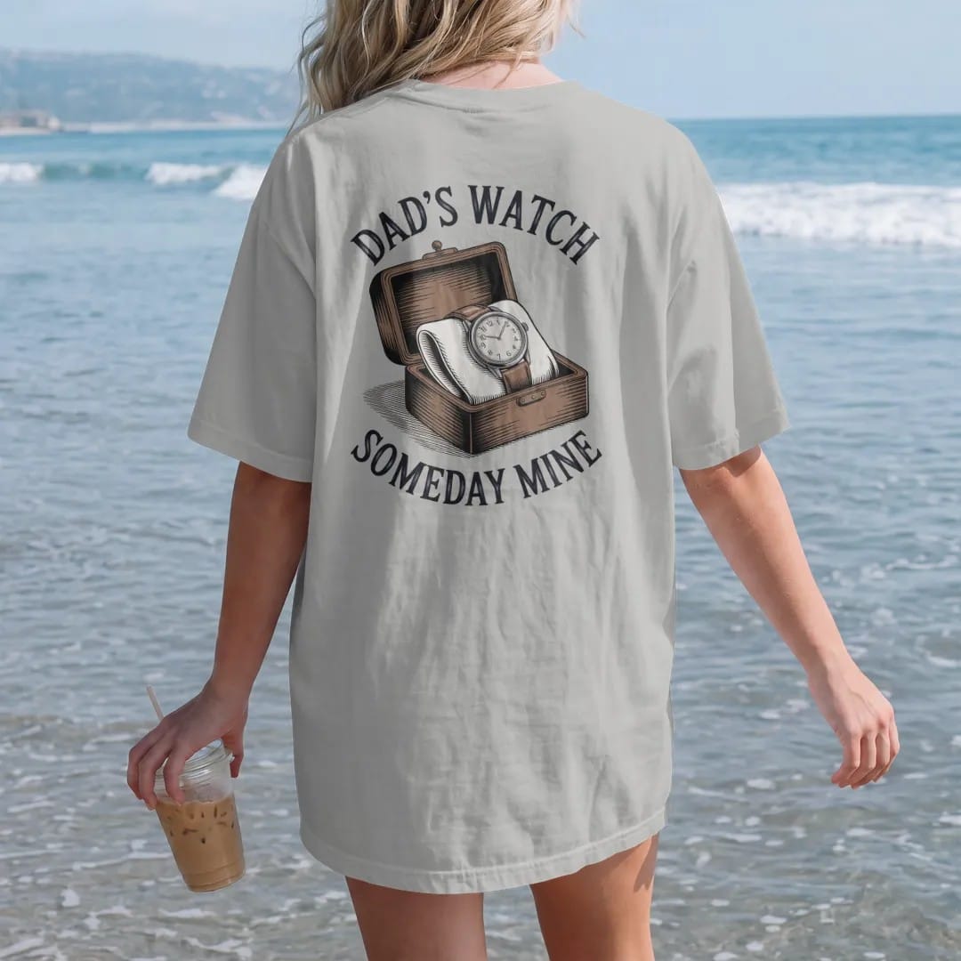Woman on the beach in a Granite tee, back view: a worn vintage watch on a leather strap in a small wooden box, under Dad's Watch and the script Someday Mine, as a fine engraving in navy and antique brass.