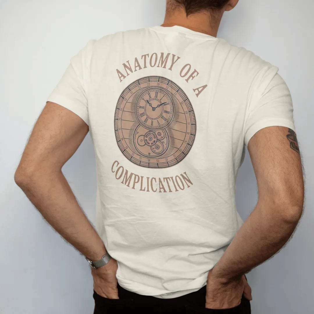 Man wearing a Ivory tee, seen from the back: a patent-style exploded watch drawing with numbered leader lines, under Anatomy Of A and Complication, as a fine engraving in navy and antique brass.