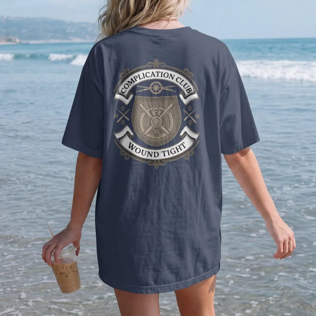 Woman on the beach in a Navy tee, back view: a shield crest with interlocking gears, a balance wheel and crossed screwdrivers under Complication Club and a Wound Tight ribbon, as a fine engraving in antique gold and cream.