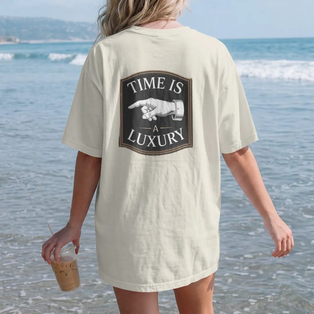 Woman on the beach in a Ivory tee, back view: a slender watch hand between a small Time Is and a large A Luxury with thin rule lines, as a fine engraving in antique gold and cream.