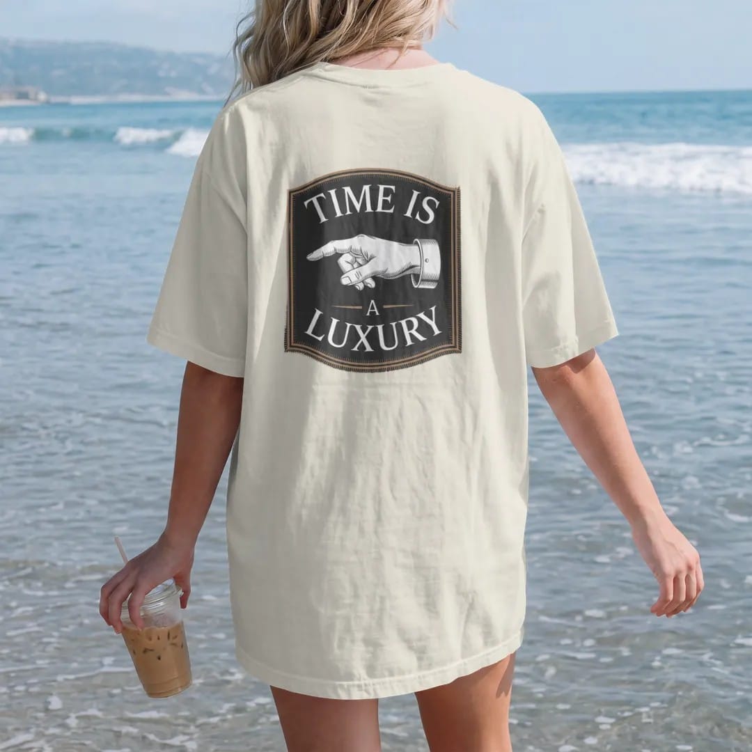 Woman on the beach in a Ivory tee, back view: a slender watch hand between a small Time Is and a large A Luxury with thin rule lines, as a fine engraving in antique gold and cream.