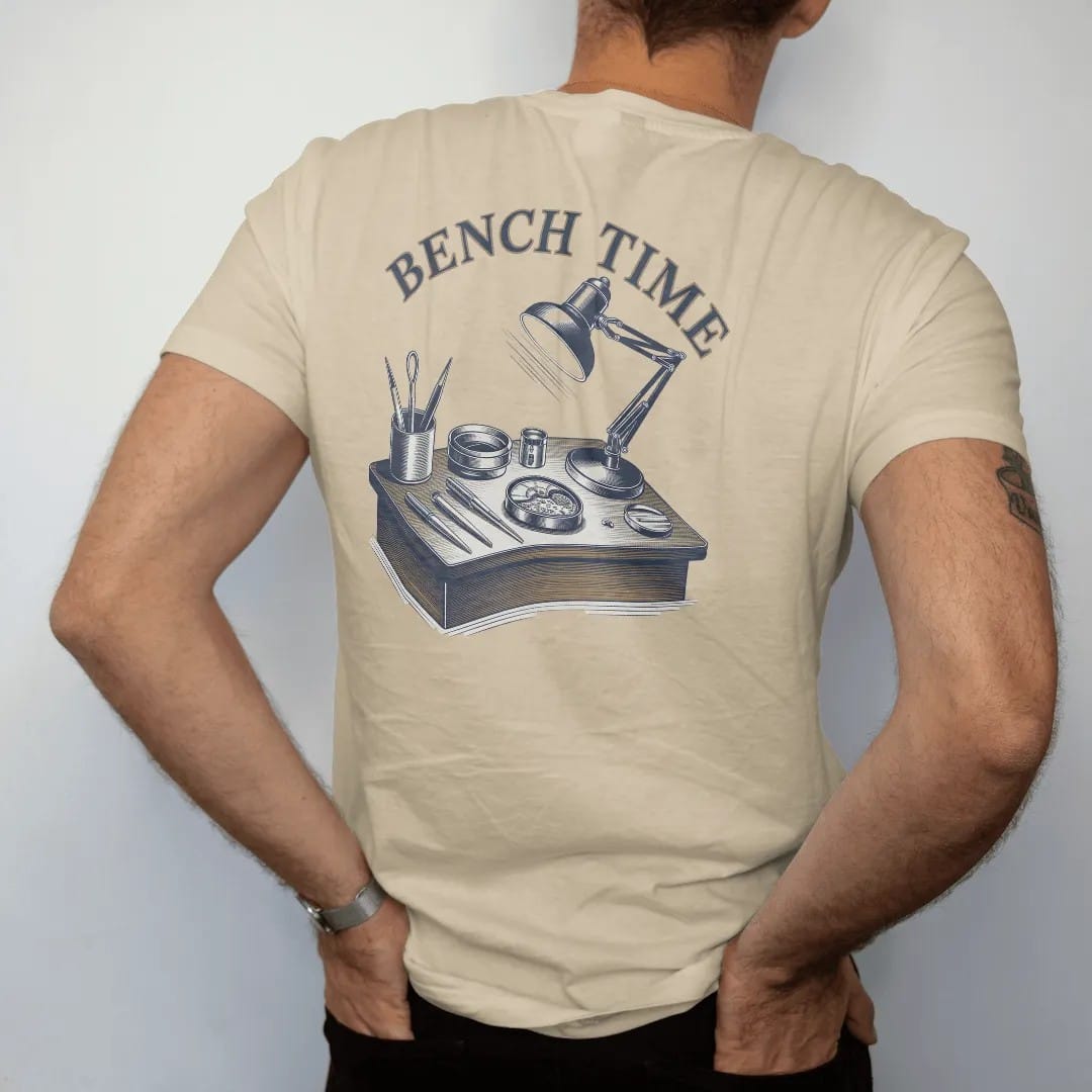 Man wearing a Sandstone tee, seen from the back: a watchmaker's bench with tweezers, screwdrivers, a loupe and an open movement under a lamp, below Bench Time, as a fine engraving in navy and antique brass.