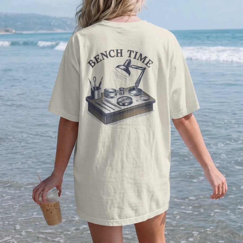 Woman on the beach in a Ivory tee, back view: a watchmaker's bench with tweezers, screwdrivers, a loupe and an open movement under a lamp, below Bench Time, as a fine engraving in navy and antique brass.