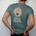 Man wearing a Blue Spruce tee, seen from the back: a watch dial with an open window revealing the balance wheel inside a small heart outline, above Open Heart, as a fine engraving in antique gold and cream.