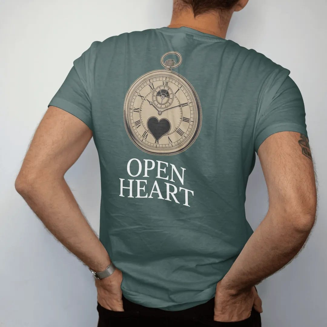 Man wearing a Blue Spruce tee, seen from the back: a watch dial with an open window revealing the balance wheel inside a small heart outline, above Open Heart, as a fine engraving in antique gold and cream.