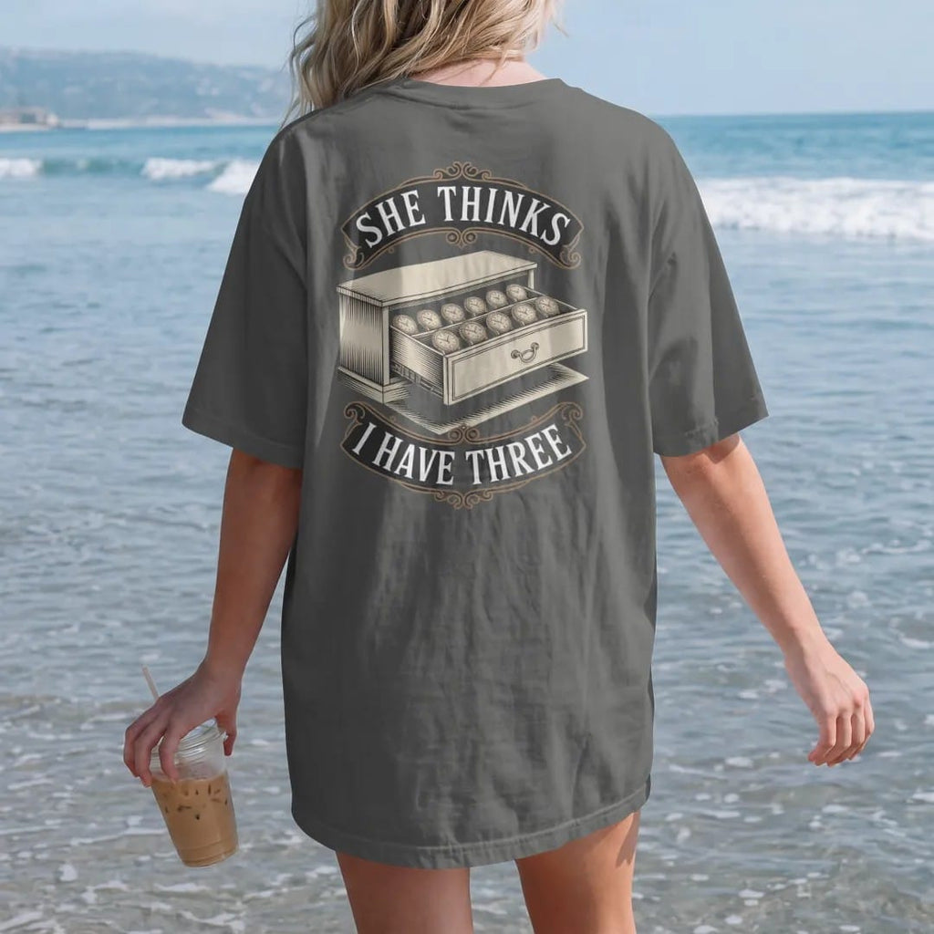 Woman on the beach in a Pepper tee, back view: a drawer open just enough to reveal a long row of hidden watches with a shushing finger icon, under She Thinks and I Have Three, as a fine engraving in antique gold and cream.